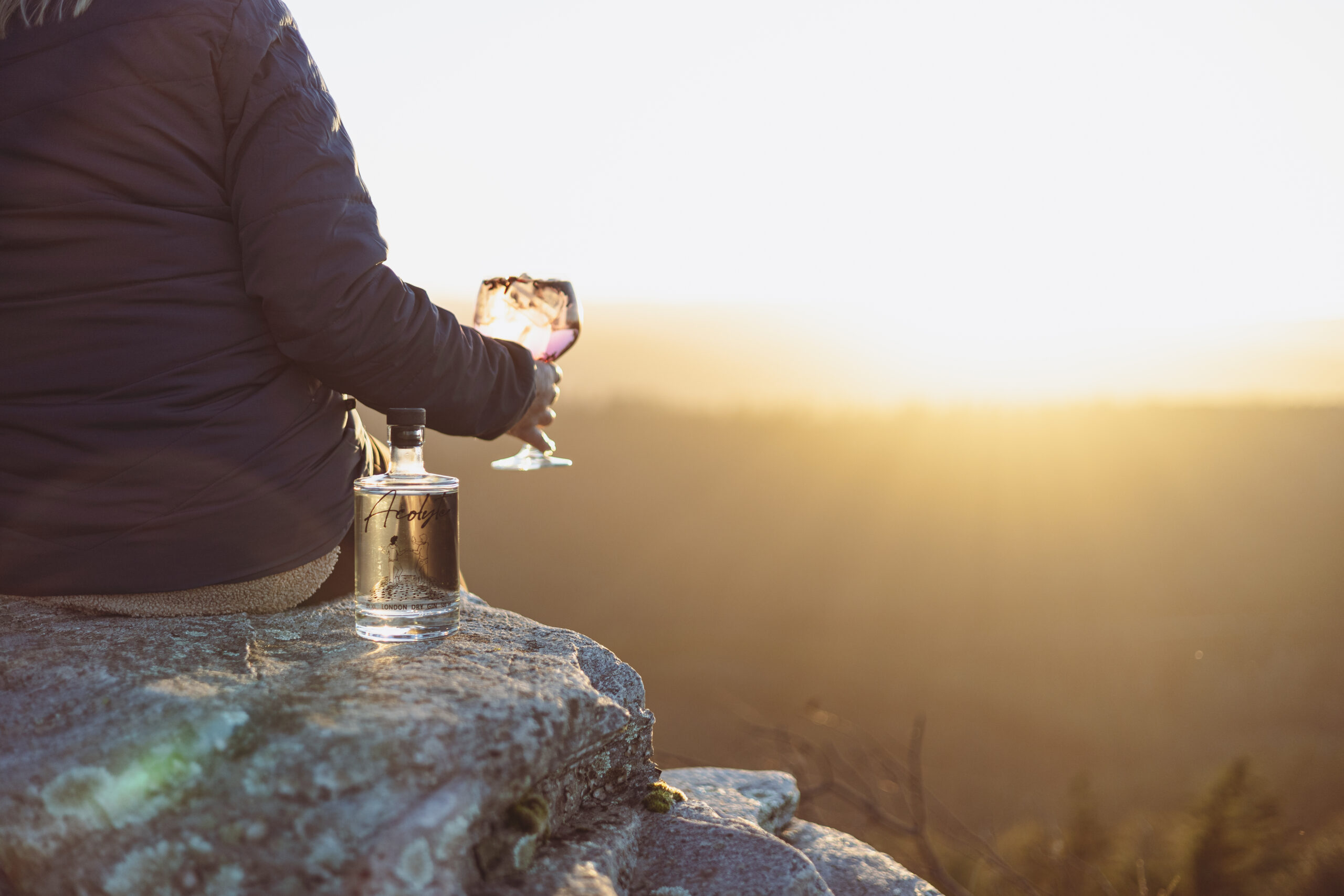 Une personne buvant un verre de Gin Acolyte préparer avec in tonic, tout en étant devant un coucher de soleil dans la nature.